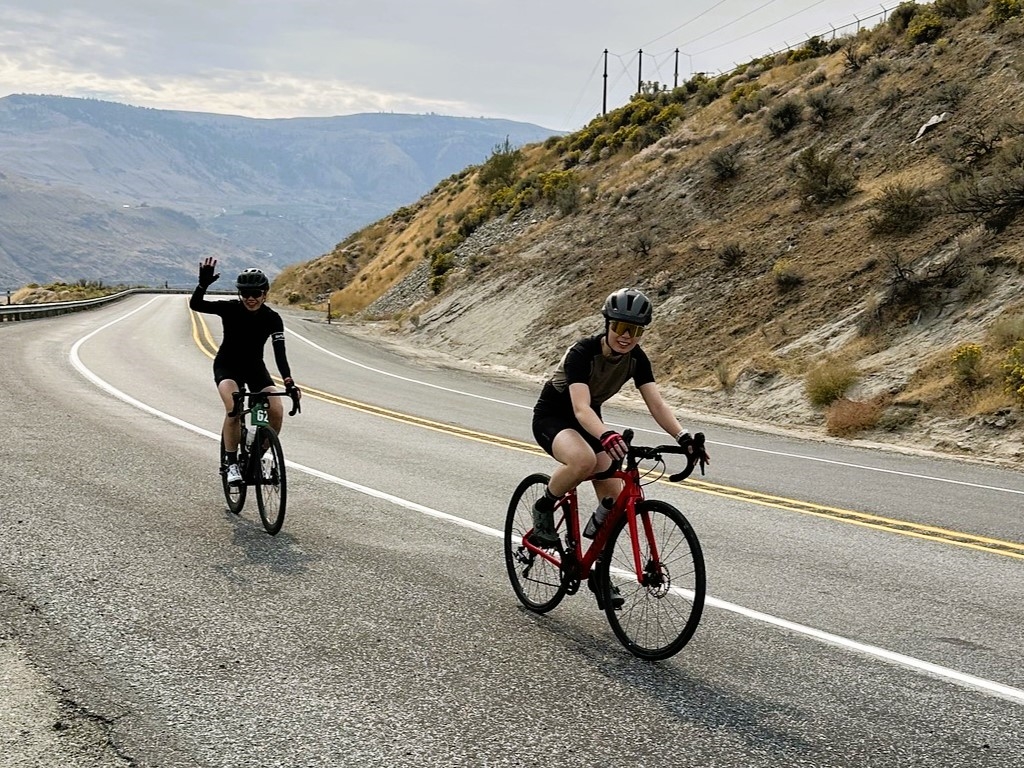 Riders smile and wave while riding up a hill on the Lake Chelan Tour.