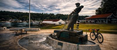 A bronze sculpture of a fisherman in a marina setting. A bicycle and a US flag in the in the background.