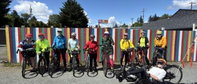 Group of bicyclists standing in front of a multi-color fence.  