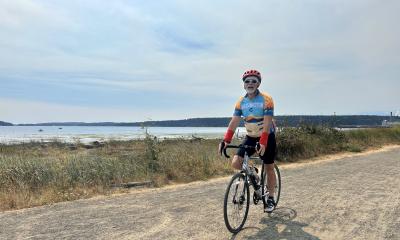 A rider cruises on a flat gravel path past Puget Sound, with marsh flora in the background.