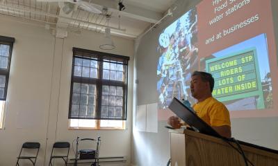 A speaker stands next to a podium in front a screen with a slide show displayed on it.