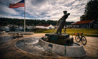 A bronze sculpture of a fisherman in a marina setting. A bicycle and a US flag in the in the background.