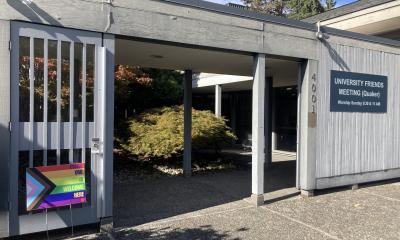 Gate leading to leafy courtyard with sign reading University Friends Meeting; a rainbow yardsign reads All Are Welcome