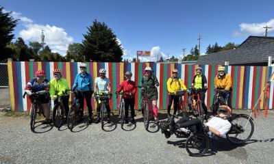 Group of bicyclists standing in front of a multi-color fence.  